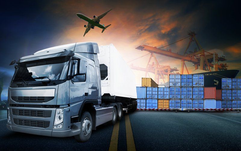 White freight truck, stacked shipping containers, and cranes at an industrial port with an airplane overhead