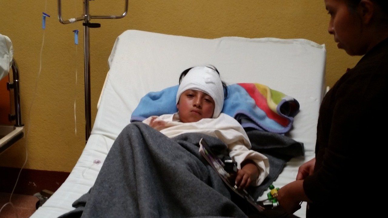 Eine Person mit Kopfverband liegt im Krankenhausbett und wird von einer Person in dunkler Kleidung betreut.