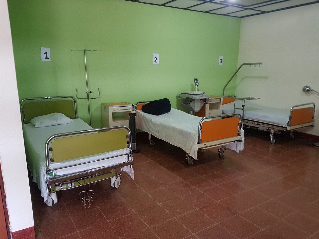 Drei leere Krankenhausbetten in einem Zimmer mit grünen Wänden und gefliestem Boden.