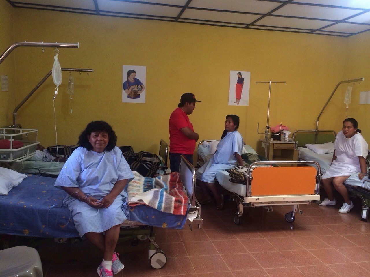 Krankenzimmer mit Patienten in Betten, einer sitzt, zwei andere stehen.