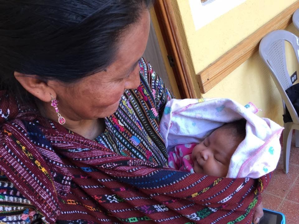 Eine Frau hält ein in ein buntes Tuch gewickeltes Baby im Arm und lächelt dem Säugling zu.