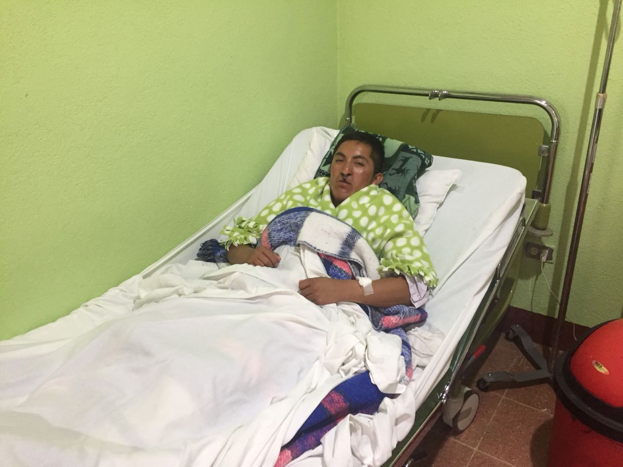 Eine Person liegt in einem Krankenhausbett, in eine Decke gehüllt. Grüne Wände, Infusionsständer und medizinische Geräte.