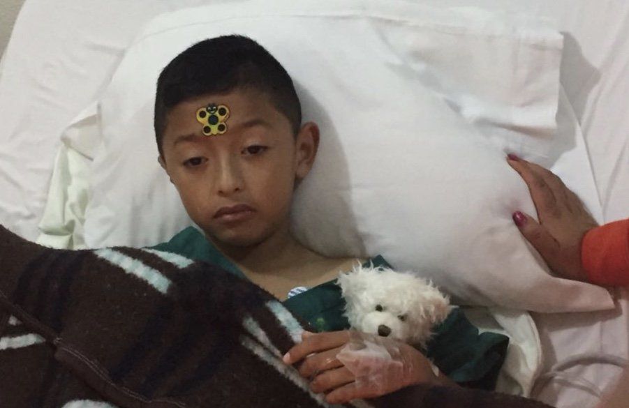 Junge im Krankenhausbett mit Teddybär, Hand am Arm, Verband auf der Stirn.
