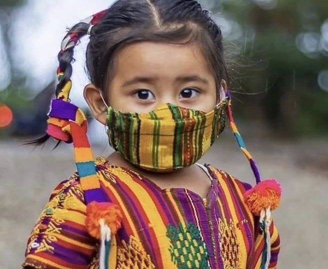 Mädchen mit gemusterter Gesichtsmaske und traditioneller, bestickter Kleidung, Zöpfen und bunten Quasten.