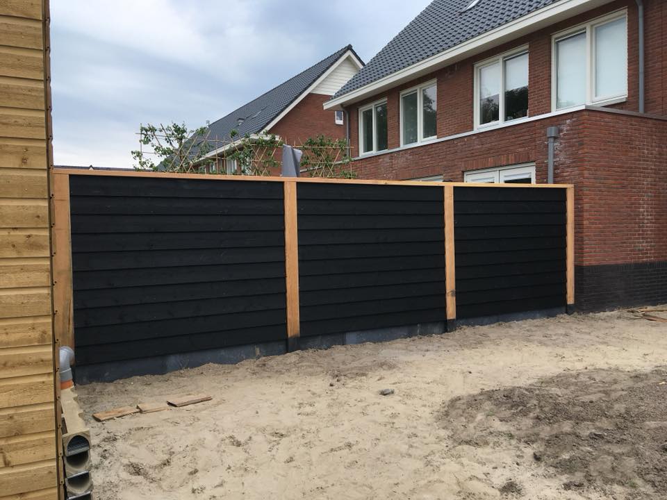Een moderne, zwarte schutting van horizontale latten scheidt een rood bakstenen huis van een zanderige, onafgewerkte achtertuin.