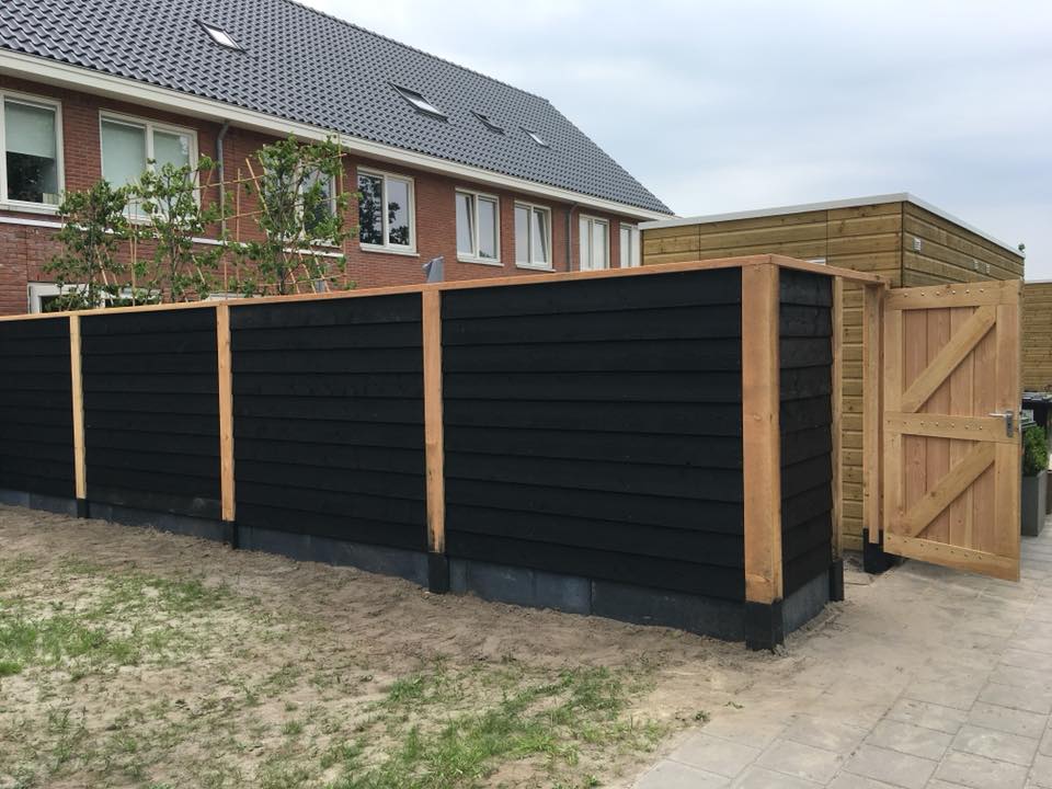 Moderne tuinafscheiding met horizontale zwarte panelen en natuurhouten palen, met een houten poort en daarachter een bakstenen huis.