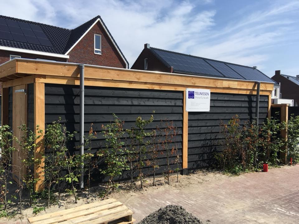 Een moderne, rechthoekige houten schuur met zwarte horizontale bekleding en een plat dak, gelegen in een woonwijk.