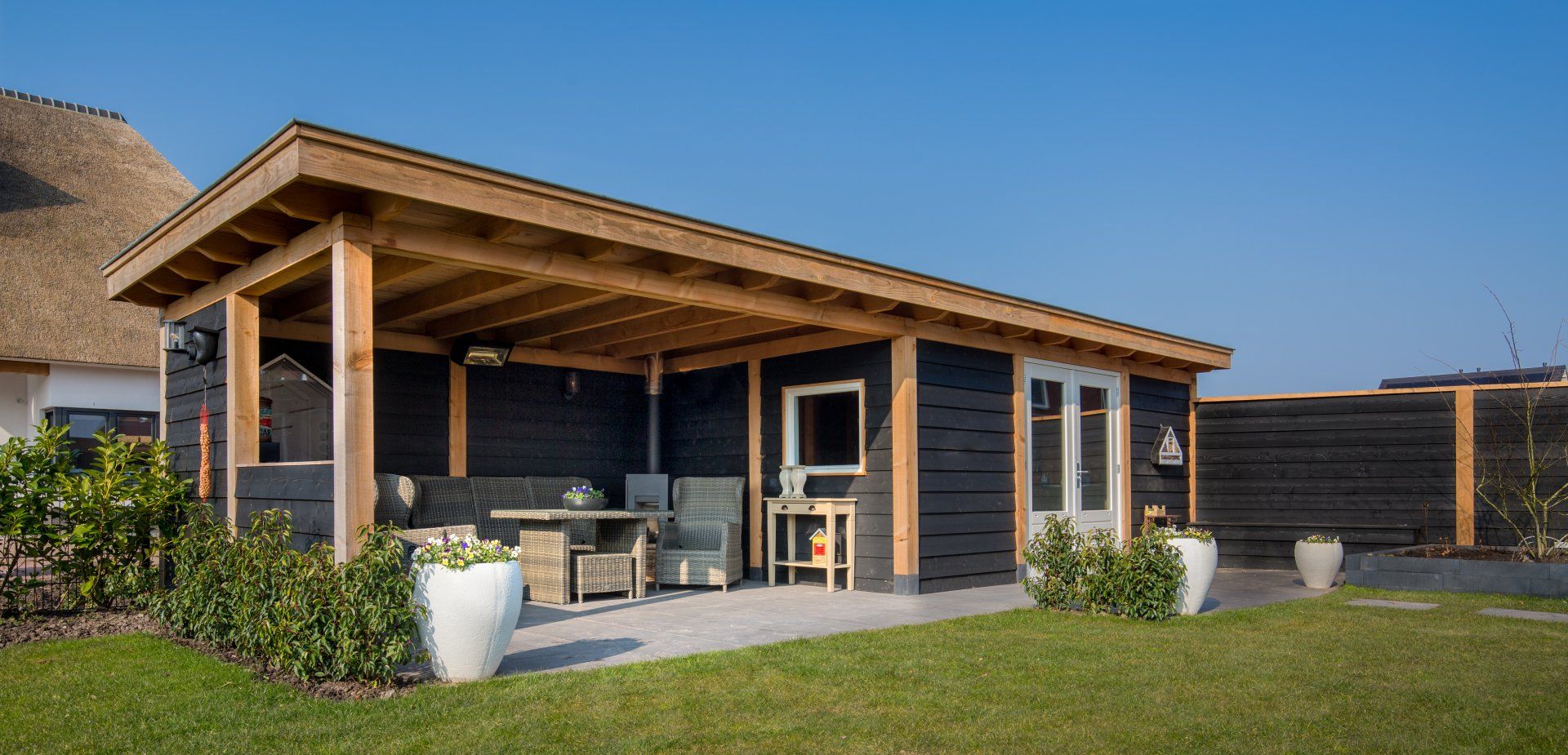 Een modern, zwart tuinhuis met een houten overkapping boven een terras, waarop op een zonnige dag tuinmeubilair staat.