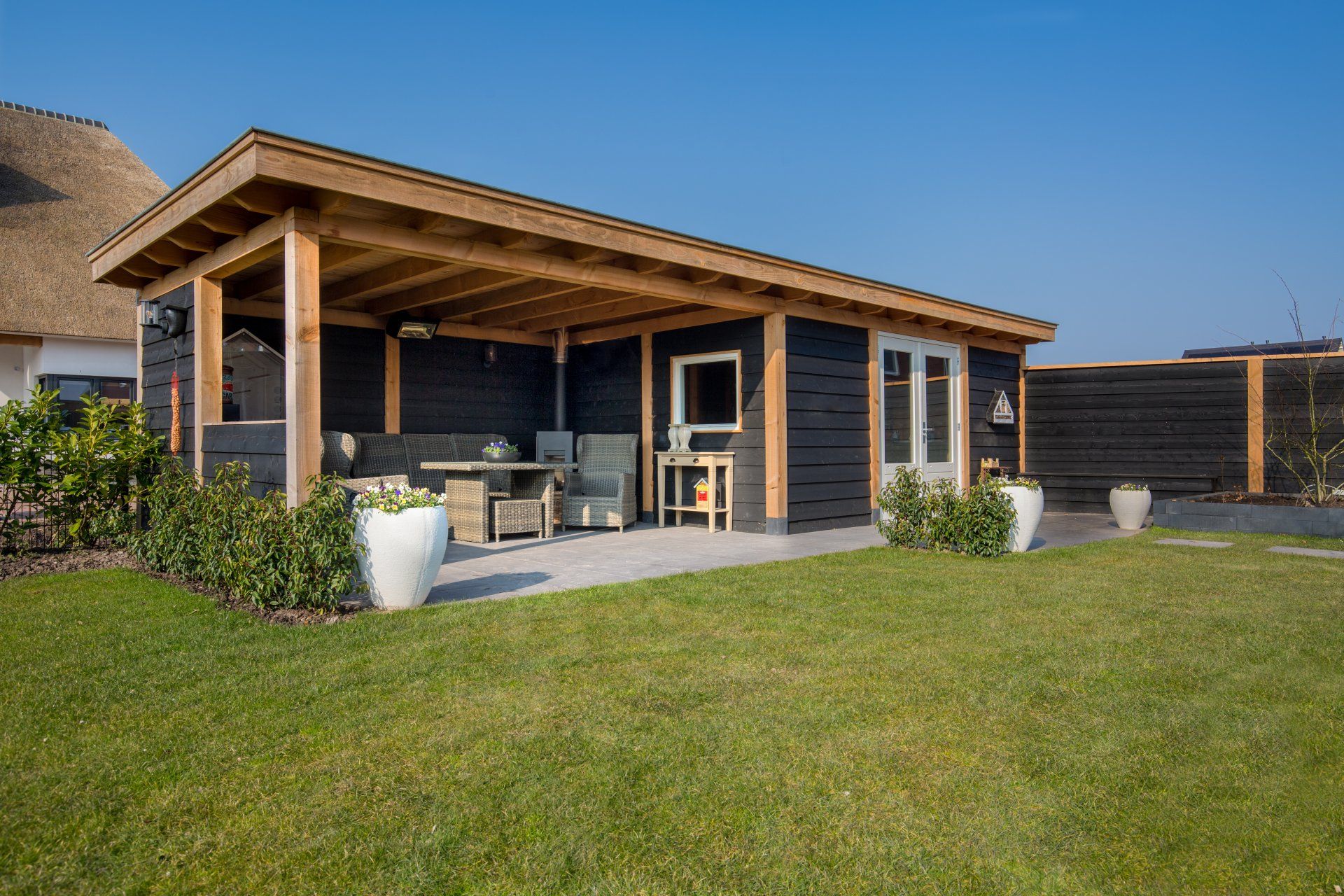 Een modern, zwart houten tuinpaviljoen met een open zitgedeelte en een glazen deur, gelegen op een heldergroen gazon.