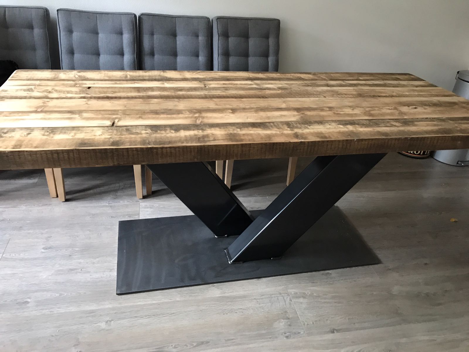 Een rustieke eettafel van houten planken met een zwart V-vormig metalen onderstel, geplaatst op een lichtgrijze houten vloer.