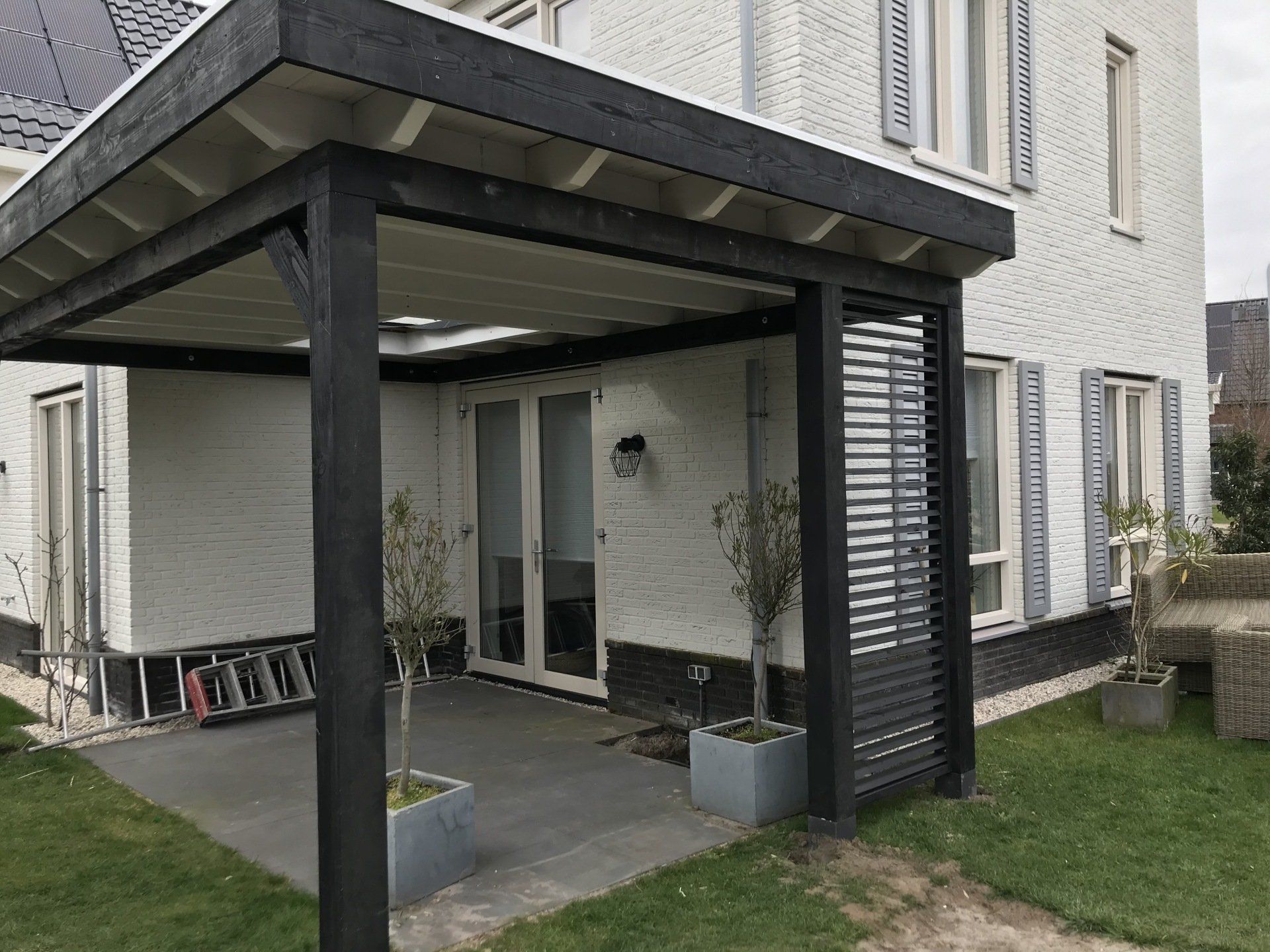 Een wit bakstenen huis met een moderne, zwarte houten terrasoverkapping en een bijpassend privacyscherm boven een betonnen terras.