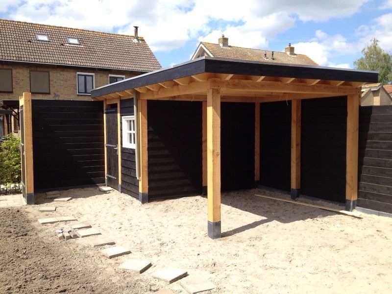 Een nieuwe, gedeeltelijk overdekte houten buitenconstructie met zwarte gevelbekleding en natuurhouten palen in een zanderige achtertuin.