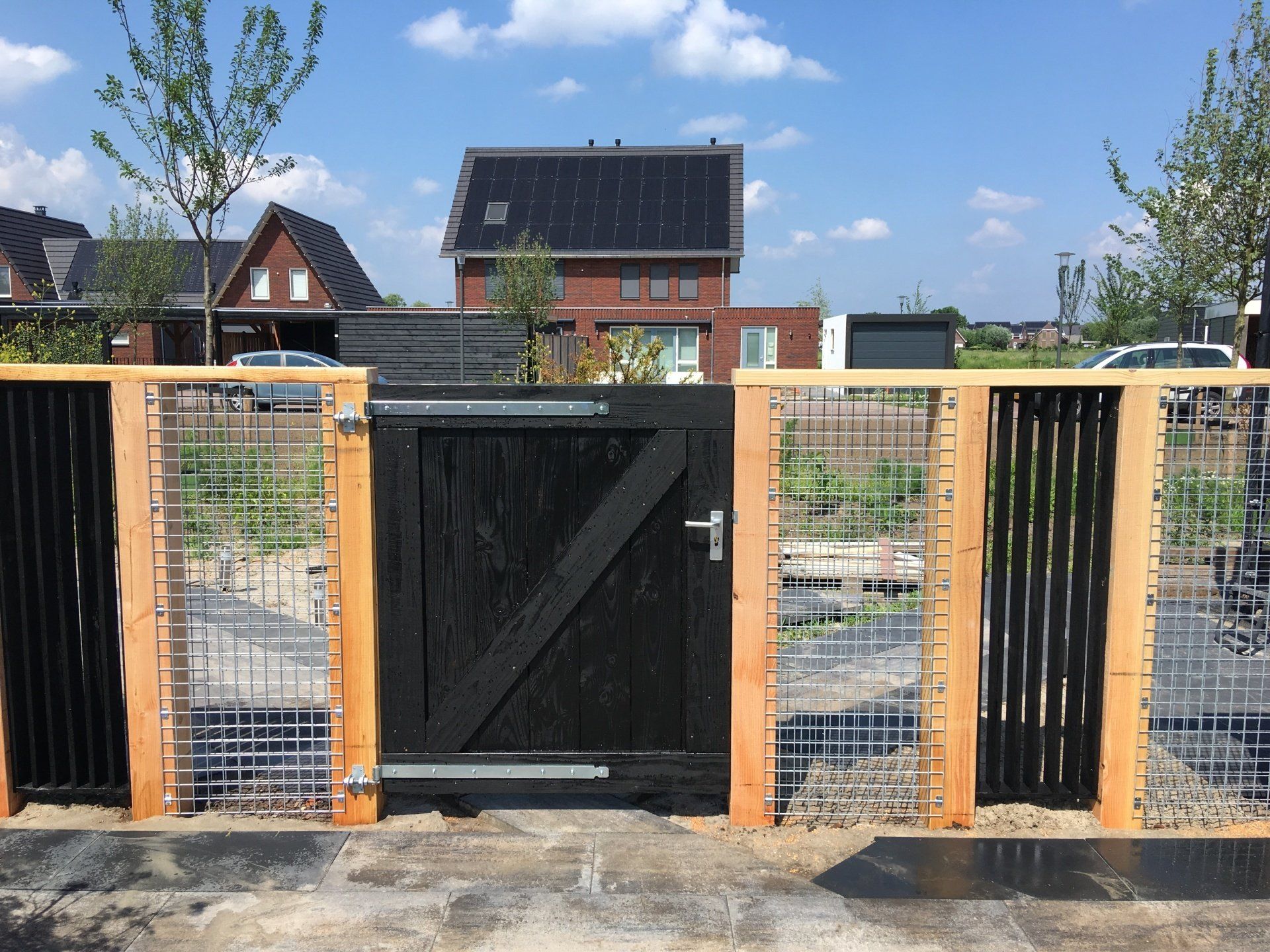 Een zwart houten hek in een omheining van lichte houten palen, metalen draad en verticale zwarte spijlen, met daarachter huizen.