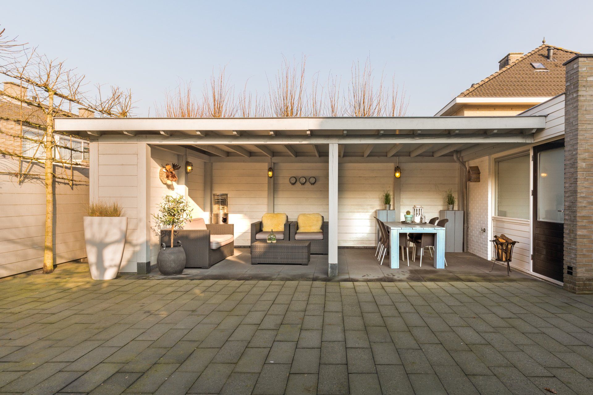 Een terras met een overdekte zit- en eethoek, voorzien van grijs tuinmeubilair, een tafel en een stenen vloer.