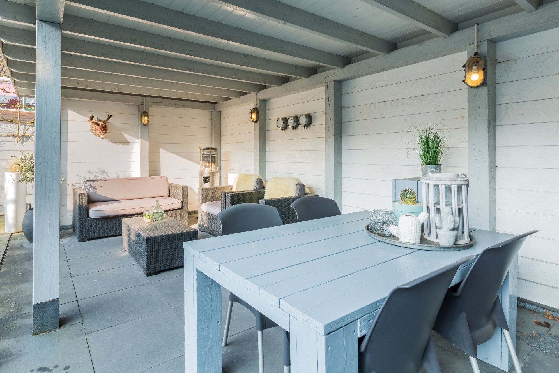 Een overdekt terras met een lichtgrijze eettafel, zitmeubels en een informele zithoek met een bank tegen witte lambrisering.
