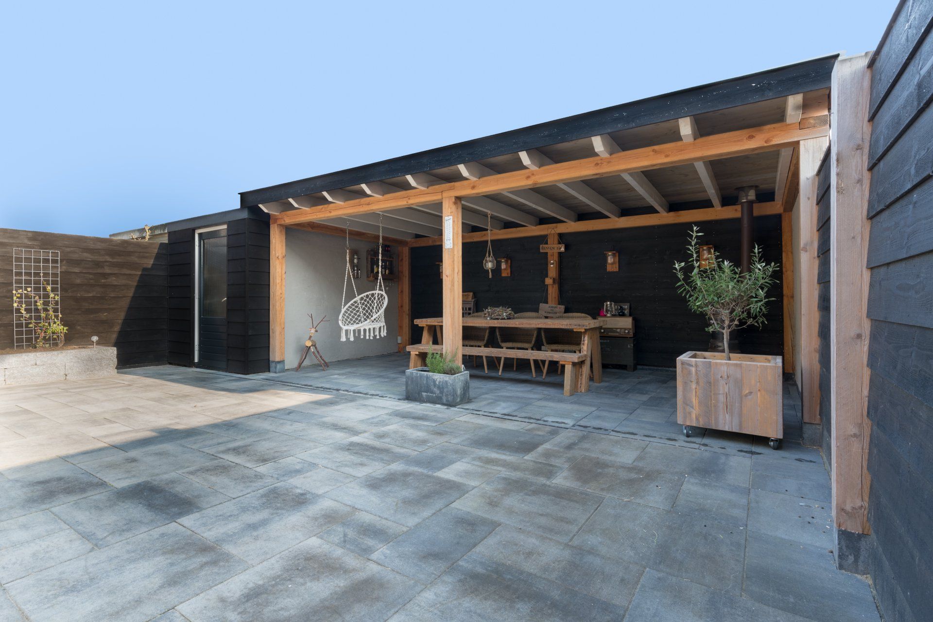 Moderne overdekte patio met een houten eethoek, schommelstoel en donkerhouten gevelbekleding, gelegen aan een grote, met stenen bestrate binnenplaats.