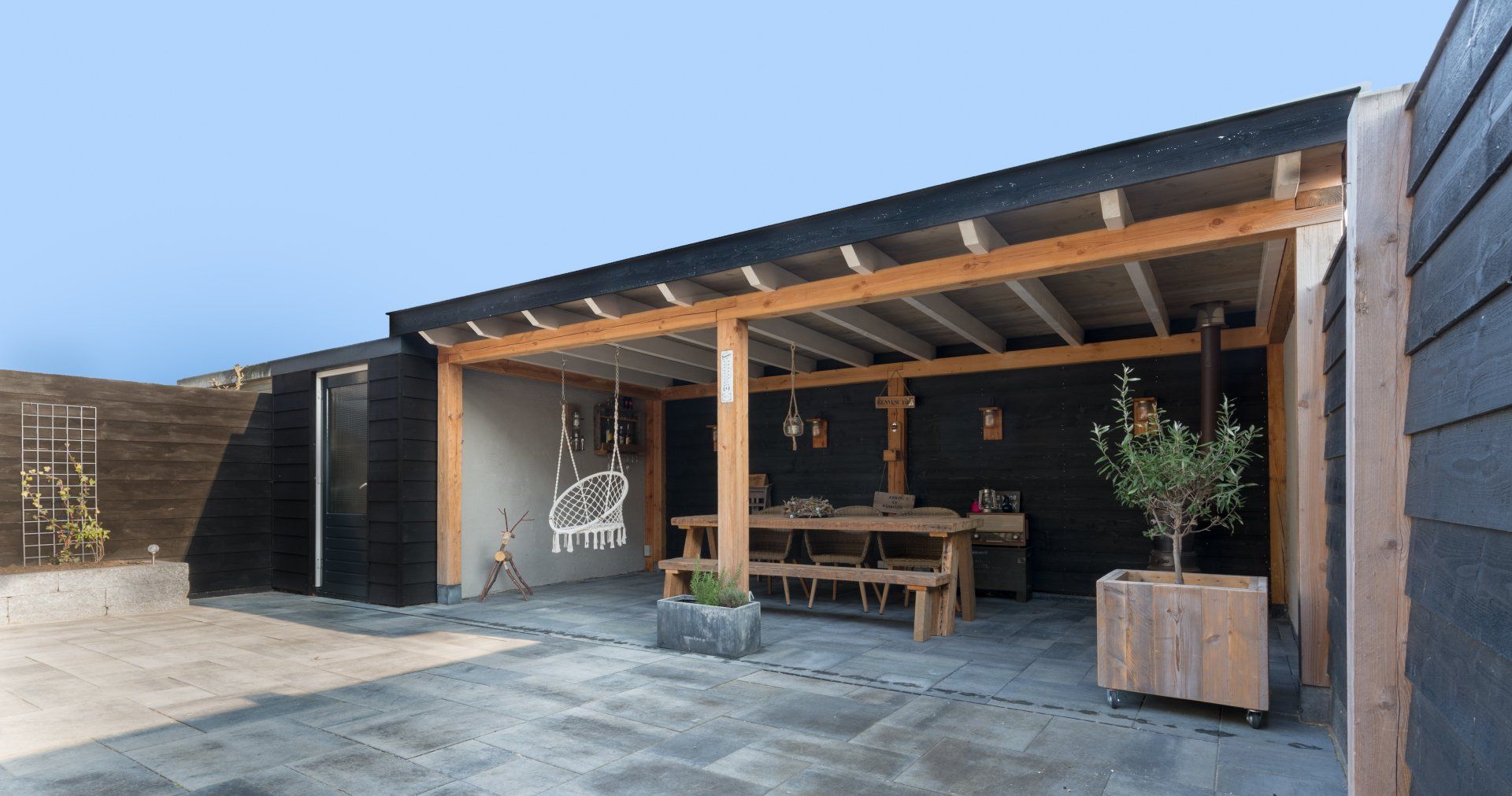 Een overdekt houten terras met een eettafel en een bank, voorzien van zwarte houten lambrisering en een stenen vloer, gelegen in een tuin.