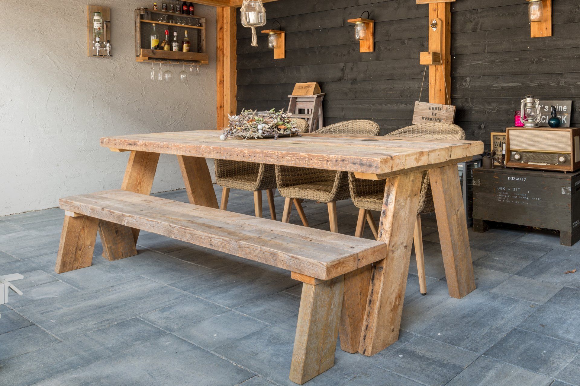 Een rustieke houten picknicktafel met een bijpassende bank op een grijze tegelvloer in een donker, sfeervol ingericht terras.