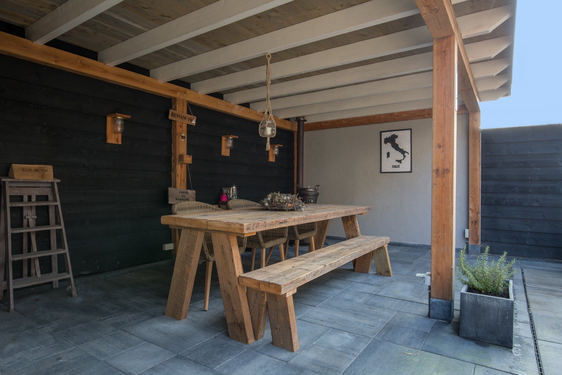 Een overdekt terras met een rustieke houten eettafel, een bijpassende bank en een klein plantje in een stenen plantenbak.