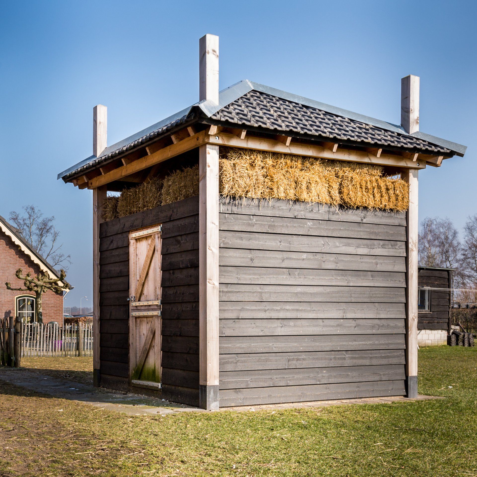 Een klein, donkerhouten schuurtje met een met stro geïsoleerd bovengedeelte en een pannendak, staat op het gras onder een blauwe hemel.