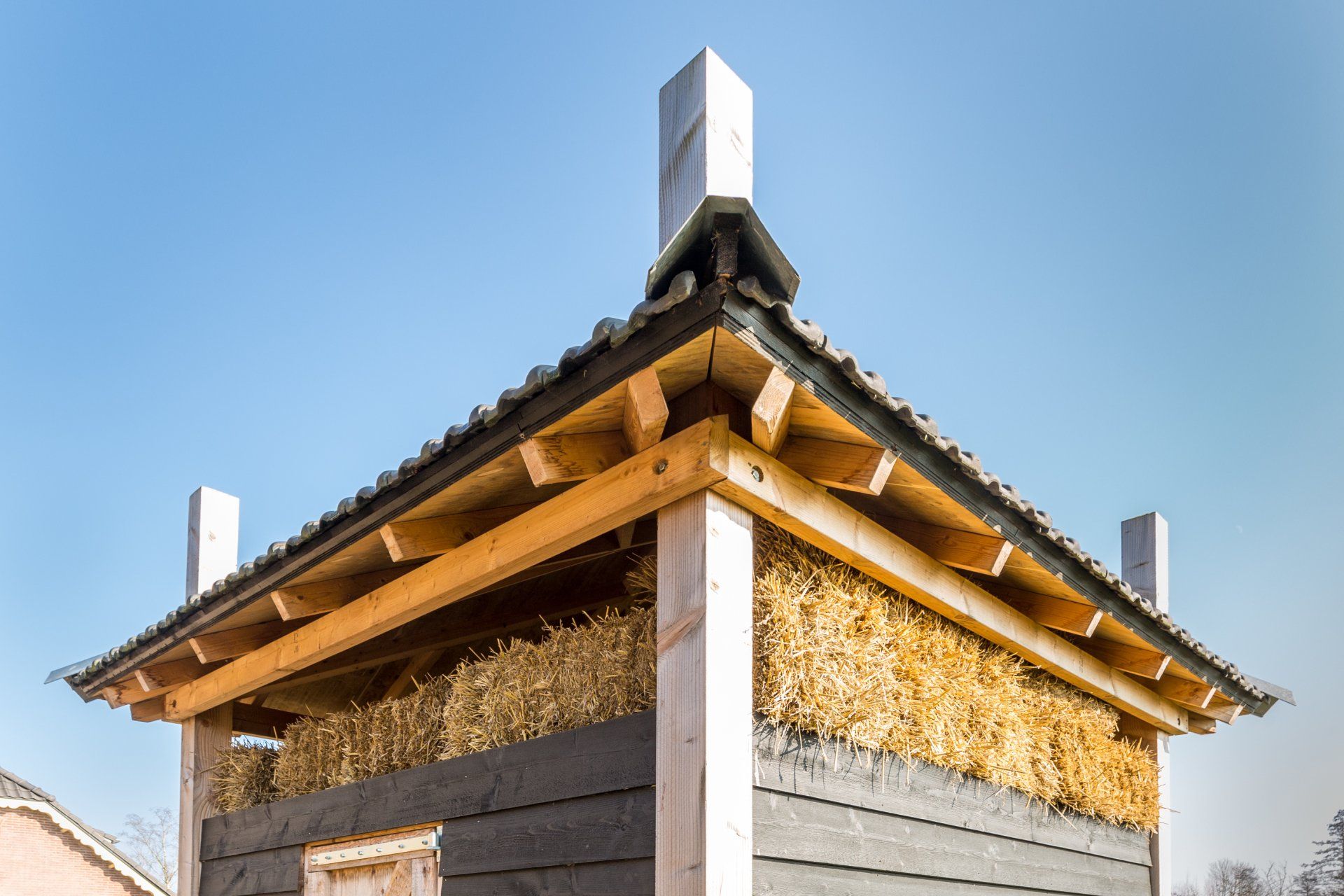 Een dakconstructie in aanbouw, met zichtbare houten balken, stro-isolatie en donkere planken tegen een blauwe lucht.