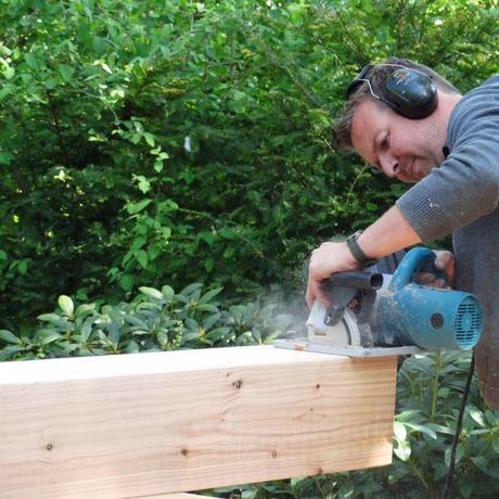 Wat een focus! Een persoon met gehoorbescherming gebruikt een blauwe cirkelzaag om een dikke houten balk door te zagen in de buitenlucht, vlakbij groen gebladerte.
