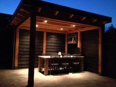 Een modern houten tuinpaviljoen met donkere lambrisering, een eettafel en ingebouwde plafondverlichting voor 's avonds.