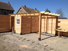 Een houten schuur en pergola in aanbouw in een zanderige tuin naast een hoge schutting die voor privacy zorgt.
