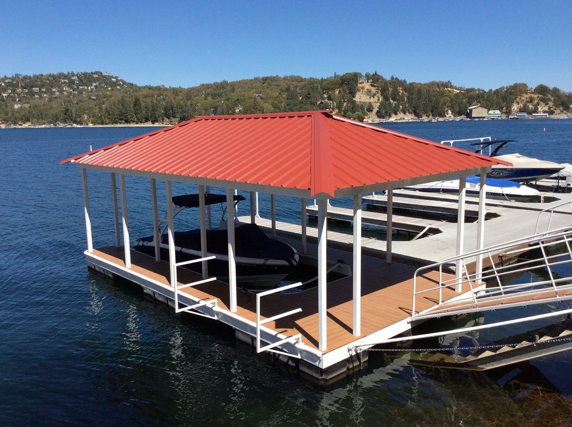 A red-roofed boat dock on a lake with boats docked. White supports and a brown deck.