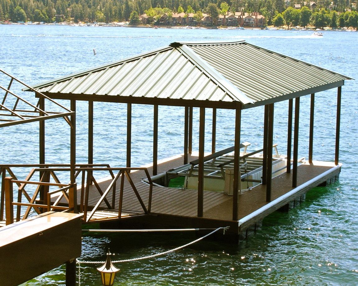 Dock with a covered boat slip on a lake. Green metal roof, brown support beams, wooden deck.