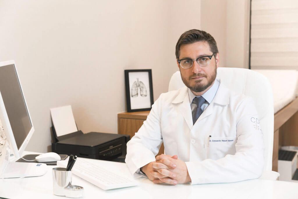 Um médico está sentado em uma mesa com as mãos cruzadas.