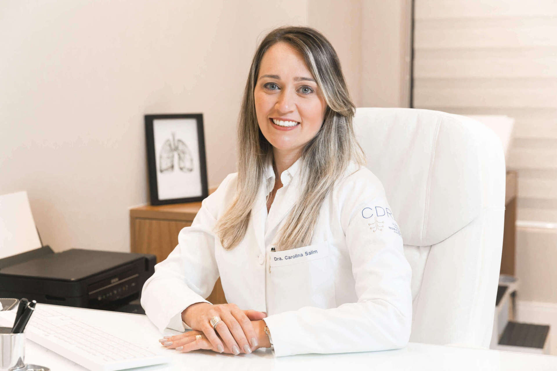 Uma mulher de jaleco branco está sentada à mesa de um escritório.