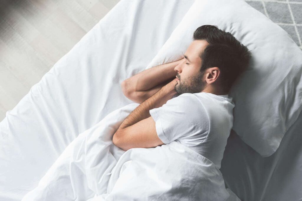 Um homem dorme em uma cama com lençóis e travesseiros brancos.