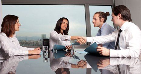 Two ladies and two men sitting round a glass table; one lady shaking hands with one of the men