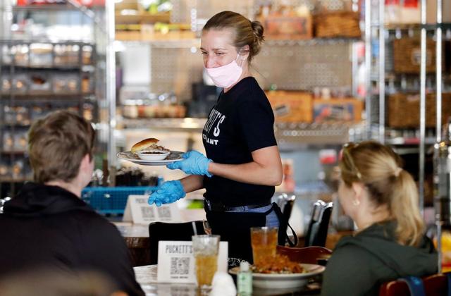 A waitress wearing a mask and gloves is serving food to a group of people sitting at a table.