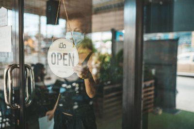 A woman wearing a mask is holding an open sign on the door of a restaurant.