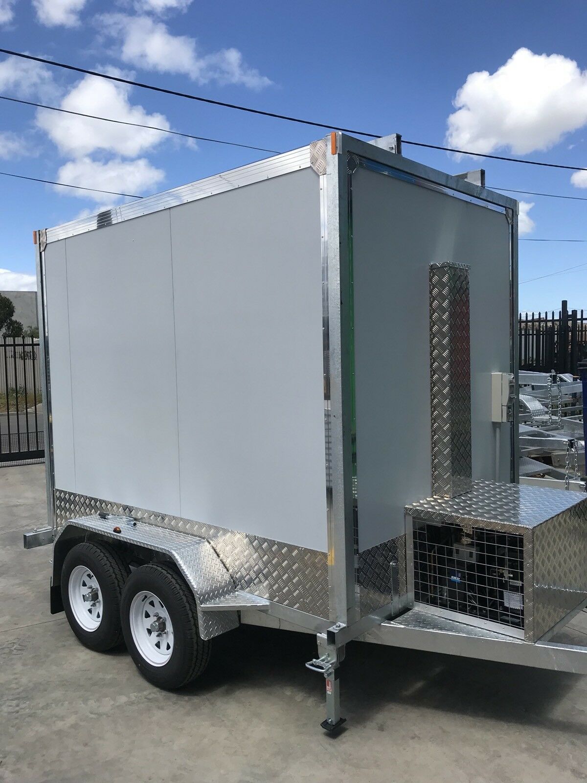 White enclosed dual-axle utility trailer with diamond-plate front platform, parked outdoors under a blue sky.