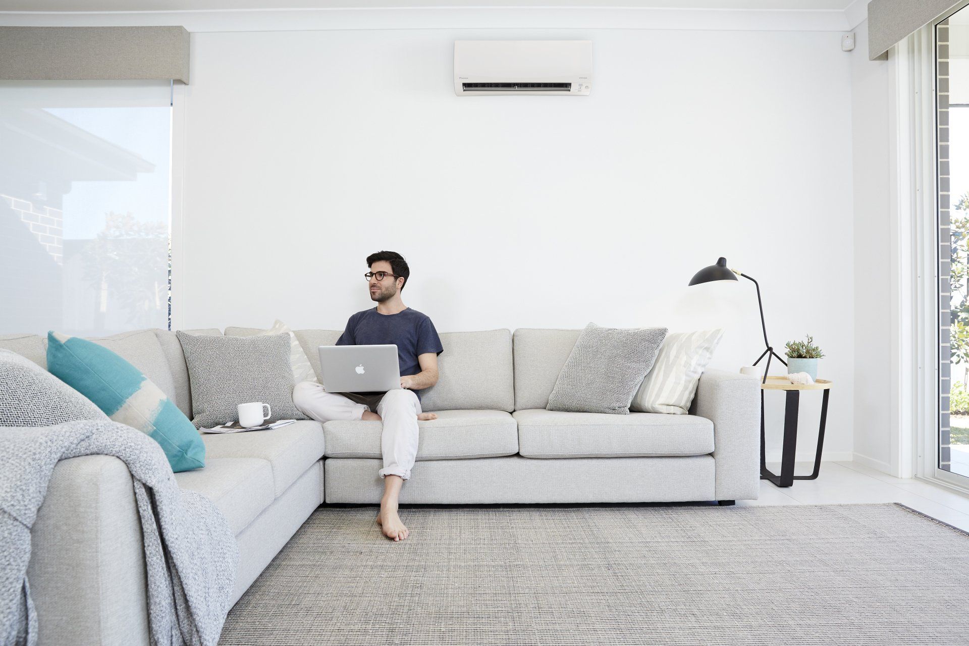 Person using a laptop on a light gray sofa in a bright living room with a floor lamp and rug.