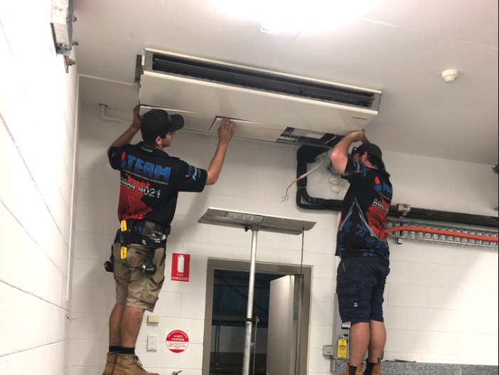 Two technicians installing a ceiling HVAC unit in a white utility room.