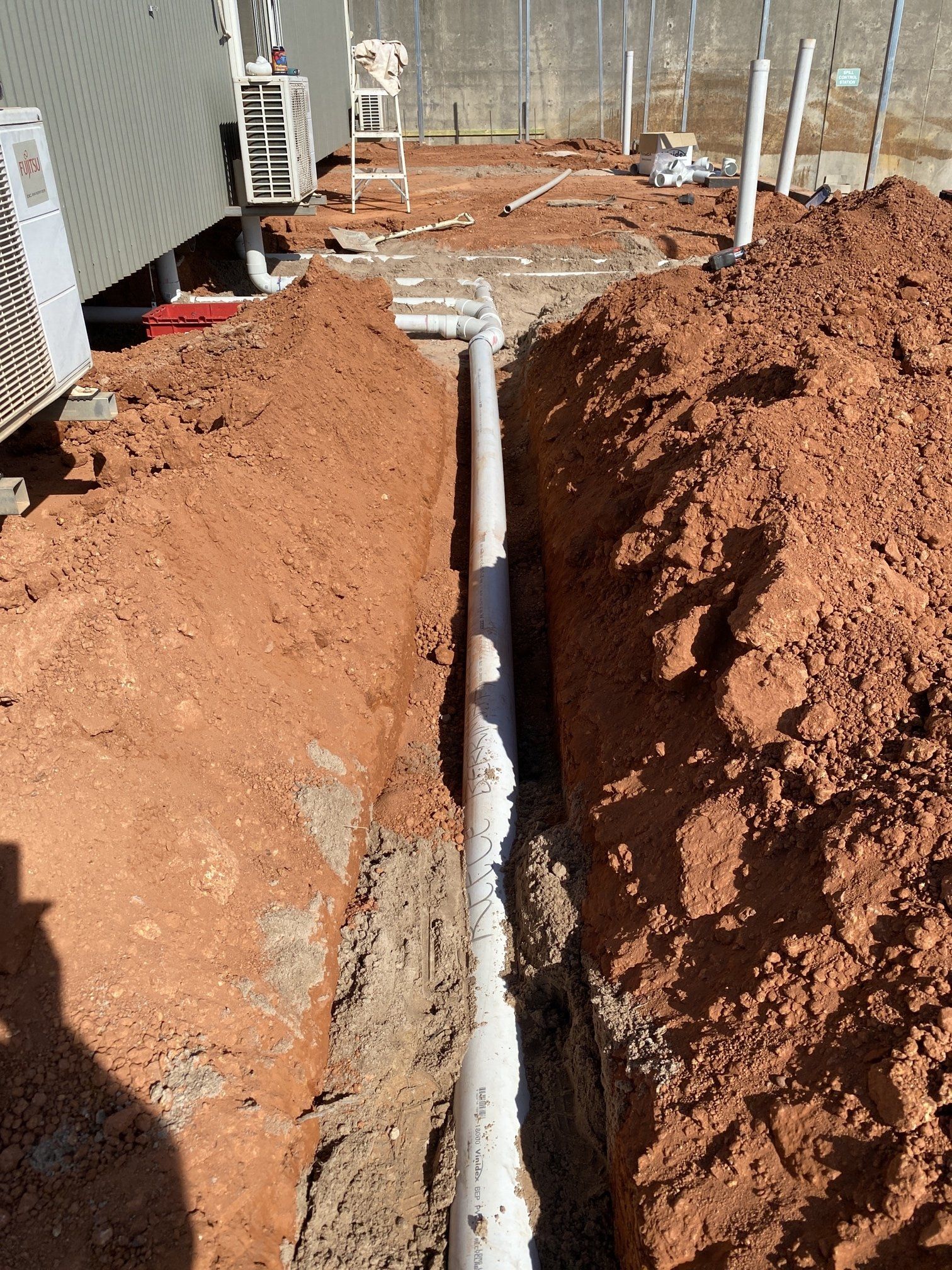 Trench dug beside a house, with exposed white pipe running between piles of red dirt.