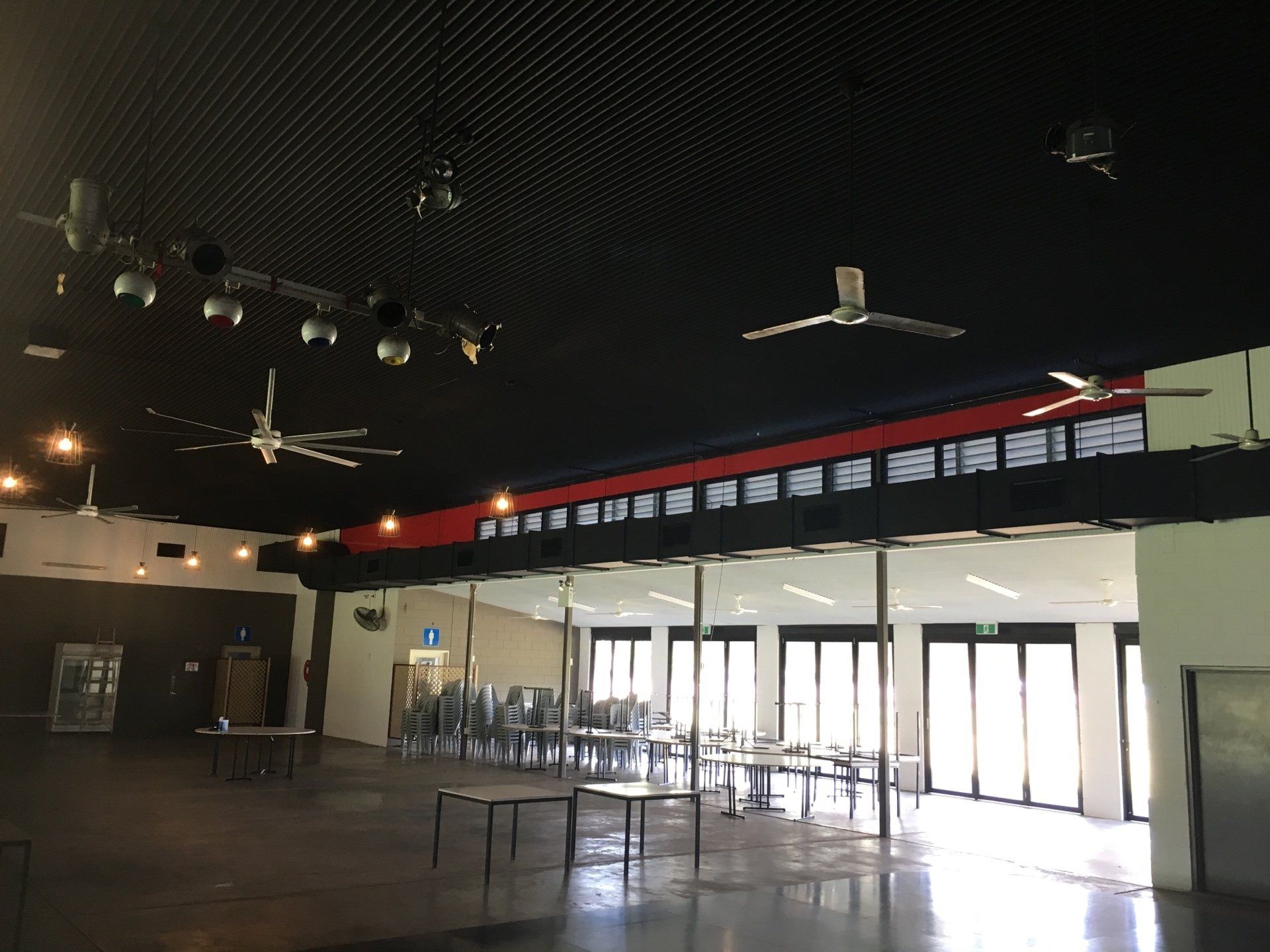 Empty event hall with high dark ceiling, hanging lights, tables, chairs, and large bright windows along one wall