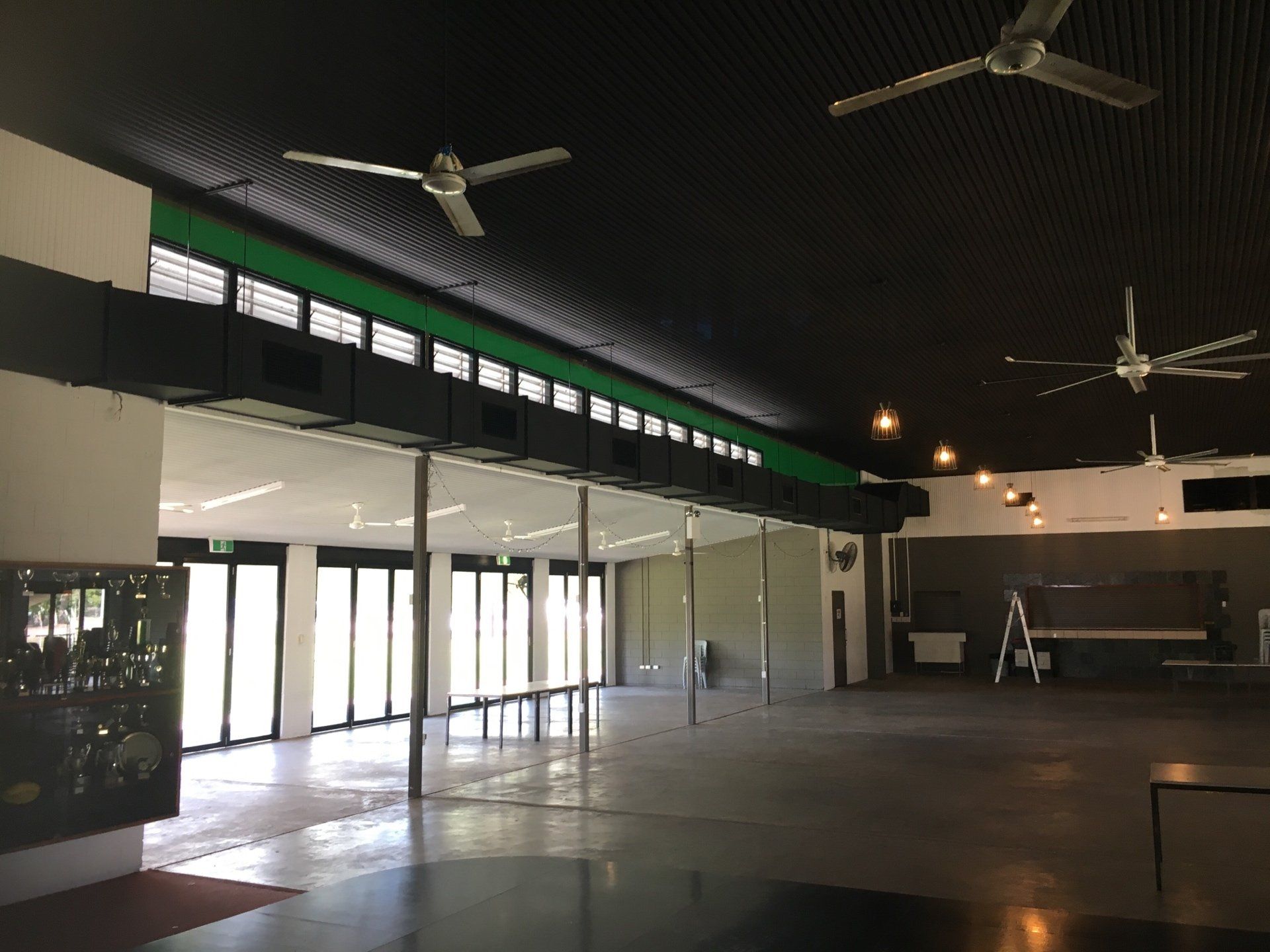 Large empty hall with high ceiling fans, glass doors, and bright light from the left side