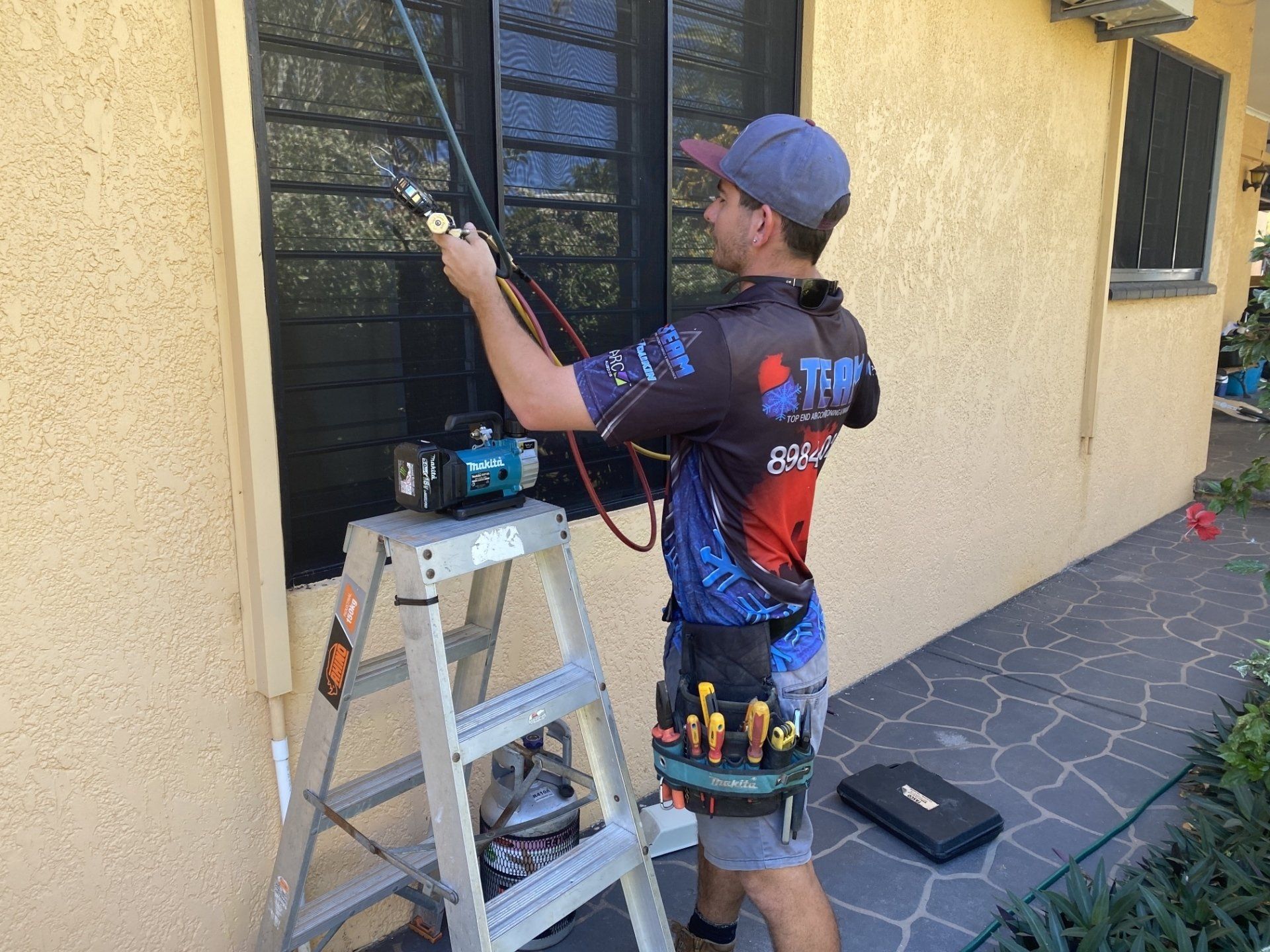 Technician on a ladder cleaning a window beside a house, with tools on a nearby step stool.
