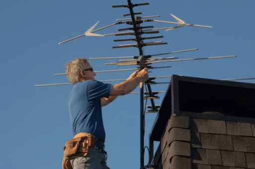 Person installing a large TV antenna on a rooftop under a clear blue sky