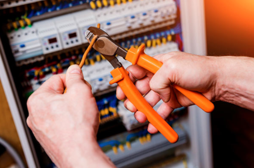 Hands using orange pliers to work on electrical wiring inside an open circuit breaker panel