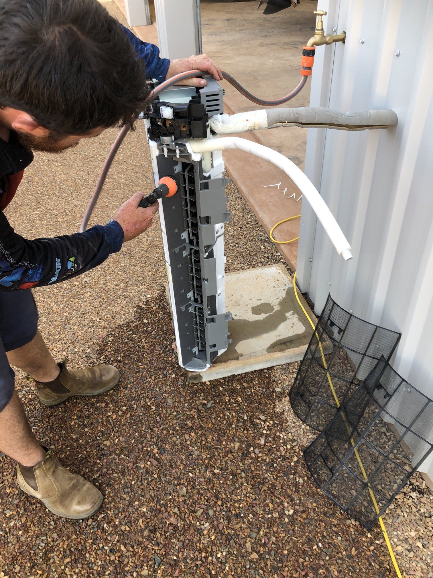 Person inspecting outdoor HVAC unit with hose and tools beside a building wall.