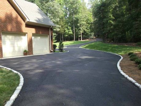 Asphalt driveway curves past a brick house with garage and into a wooded area. Edges trimmed with stones and grass.