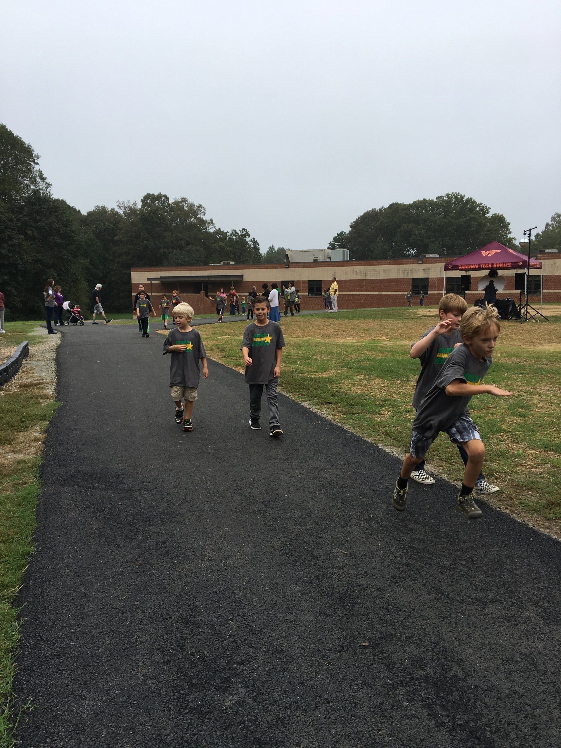 Three children running/walking on a paved path in front of a school building. One child is running ahead.