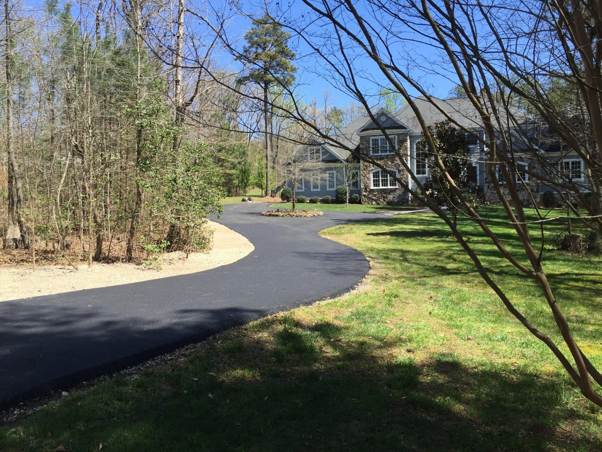 Asphalt driveway curves toward a house with gray siding, surrounded by trees and grass on a sunny day.