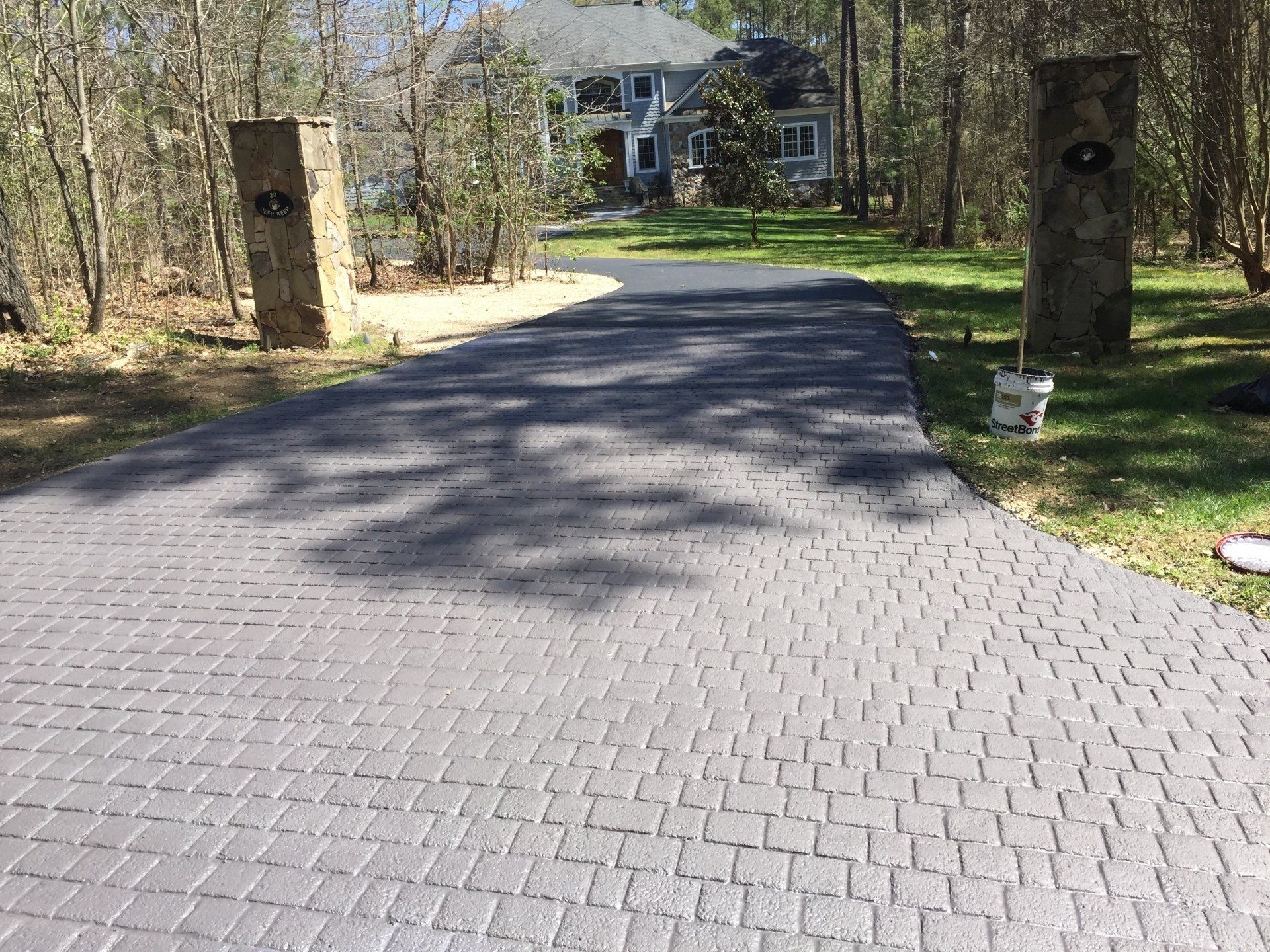 Gray brick driveway leading to a house, flanked by stone pillars, trees, and grass.